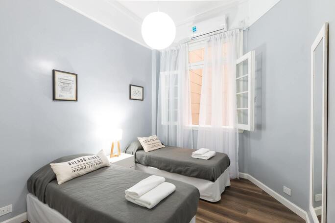 Une chambre avec deux lits simples, deux tableaux au mur et une fenêtre avec des rideaux.